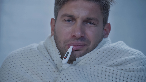 upset diseased man in blanket  while measuring temperature with thermometer in mouth