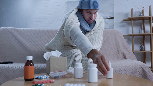 ill man in warm blanket and hat reaching nasal spray while sitting on sofa near medicaments