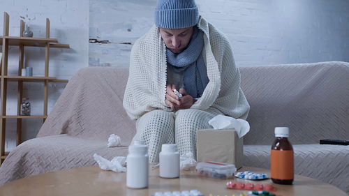 sick man in warm hat, scarf and blanket holding nasal spray near table with medication and paper napkins