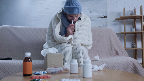 diseased man in warm blanket and beanie using nasal spray while suffering from runny nose near table with medication