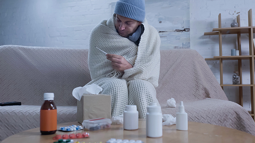 ill man in warm hat and blanket looking at thermometer near table with medication and paper napkins