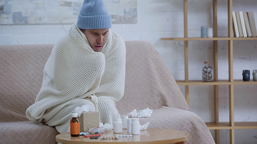 sick man sitting on couch in warm blanket and hat near paper napkins and medicaments on table