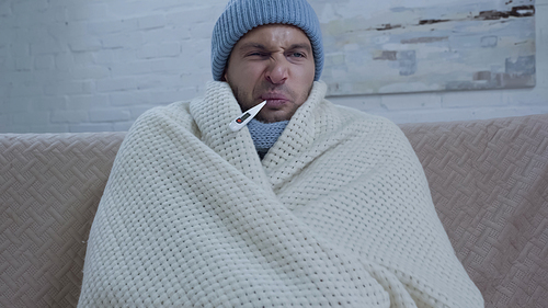 displeased sick man in warm hat and blanket measuring temperature with thermometer in mouth