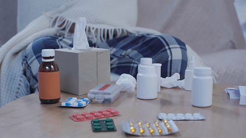 cropped view of sick man sitting on couch under warm blanket near table with medicaments and paper napkins
