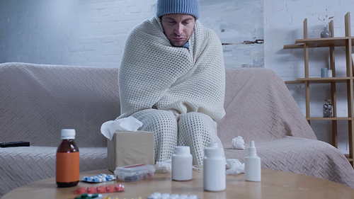 diseased man in warm blanket and hat sitting with closed eyes near table with medication and paper napkins