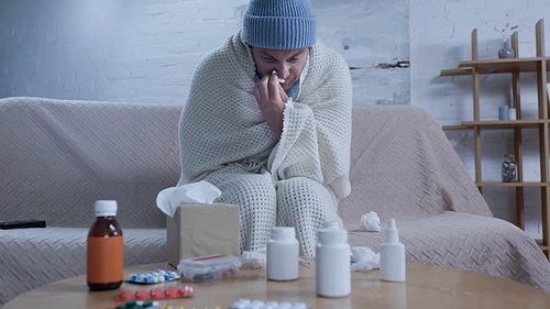 man in warm blanket and beanie suffering from runny nose near table with medication