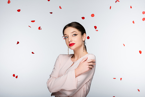 young and dreamy woman with bright makeup near falling rose petals on grey