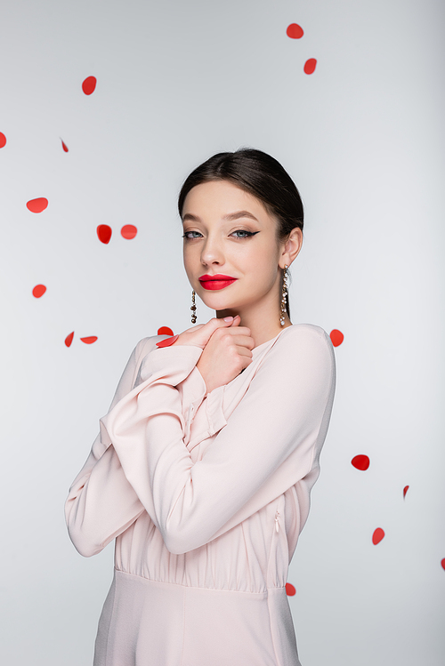 young and pleased woman with bright makeup near falling rose petals on grey