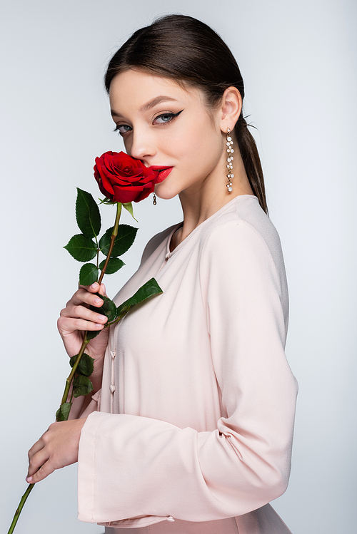 brunette woman in earrings and blouse smelling red rose isolated on grey