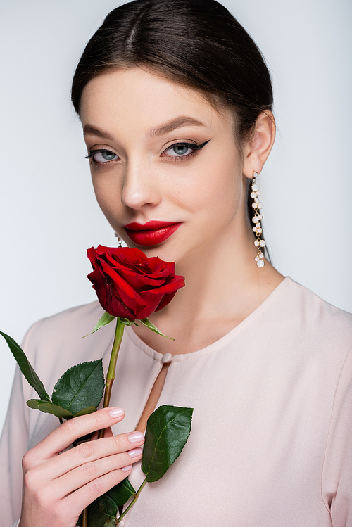 brunette young woman in earrings and blouse holding red rose isolated on grey