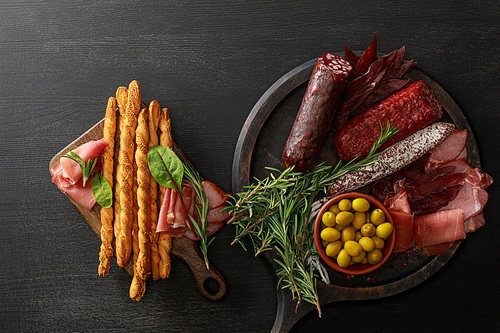 top view of delicious meat platters served with olives, breadsticks and herbs on boards on wooden black table