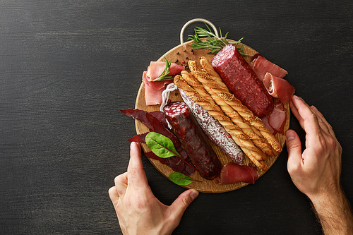 cropped view of man holding delicious meat platter served with breadsticks and herbs on board on wooden black table