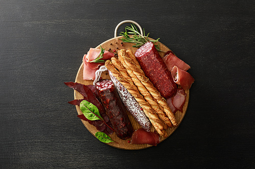top view of delicious meat platter served with breadsticks and herbs on board on wooden black table