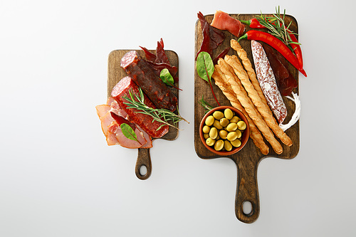 top view of delicious meat platters served with breadsticks, olives, chili peppers and herbs on boards isolated on white