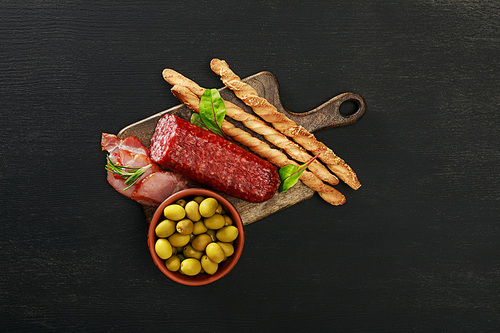 top view of delicious meat platter served with olives and grissini on board on black surface