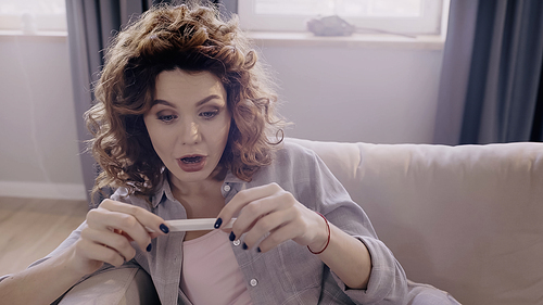 Shocked woman looking at pregnancy test on couch at home