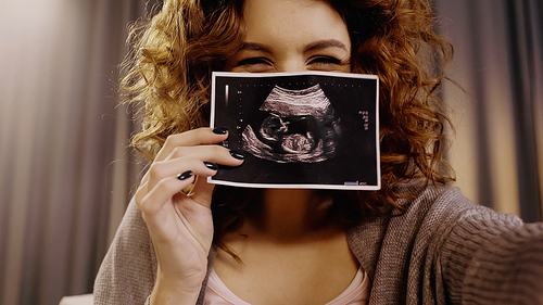 Curly woman covering face with ultrasound scan of baby at home