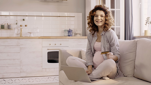 Pregnant woman holding credit card and smiling at camera near laptop on couch