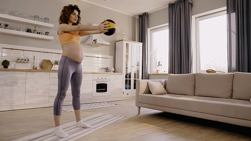 Curly pregnant sportswoman exercising with slam ball on fitness mat at home