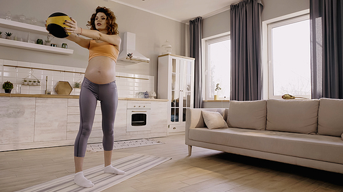 Pregnant sportswoman holding slam ball while training on fitness mat at home