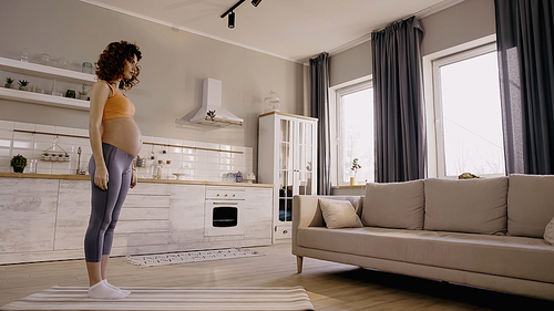 Side view of young pregnant sportswoman standing on fitness mat at home