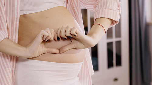 Cropped view of pregnant woman showing heart sign near belly at home