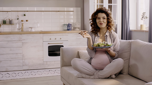 Happy pregnant woman holding plate with fresh salad and fork on couch