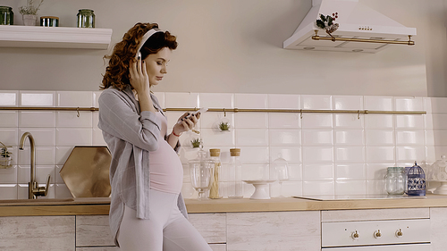 Young pregnant woman listening music in headphones and holding smartphone in kitchen