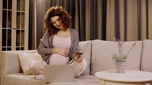 Young pregnant woman touching belly and holding credit card near laptop on couch at home