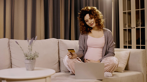 Pregnant woman holding credit card near laptop and blurred plant on couch at home