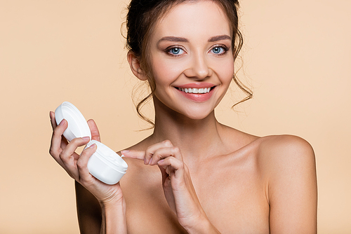 Smiling woman with naked shoulders holding cosmetic cream and  isolated on beige