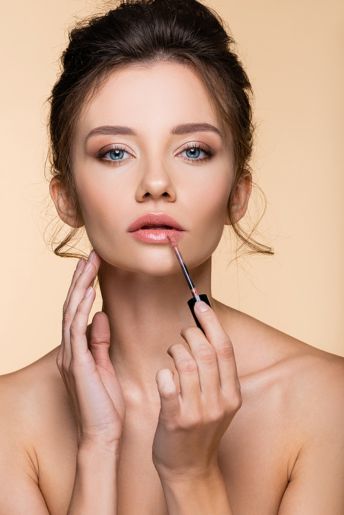 Young model with naked shoulders applying lip gloss and touching face isolated on beige
