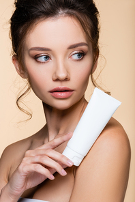 Young model holding cosmetic lotion near naked shoulder isolated on beige