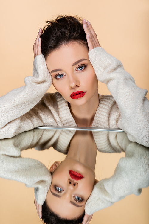 Stylish brunette model touching hair near mirror isolated on beige