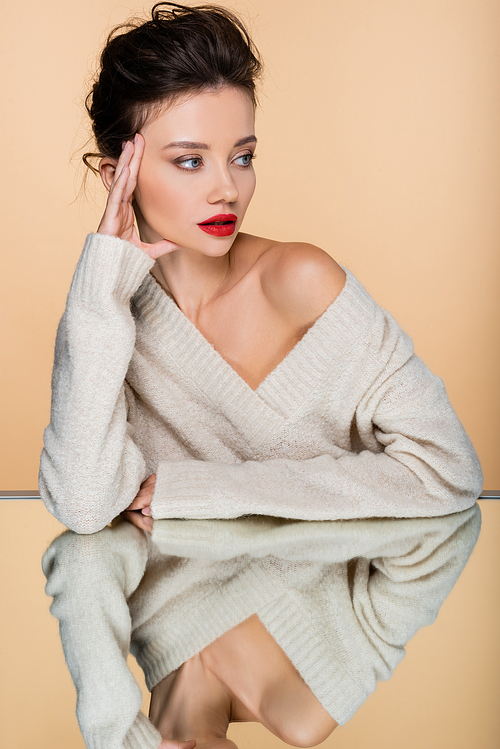 Trendy model in sweater with naked shoulder looking away near mirror isolated on beige