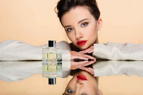 Stylish young woman in warm sweater posing near perfume bottle on mirror isolated on beige
