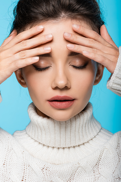 Portrait of young brunette woman in sweater touching forehead isolated on blue