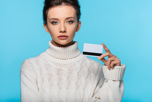 Young woman in knitted sweater holding credit card isolated on blue