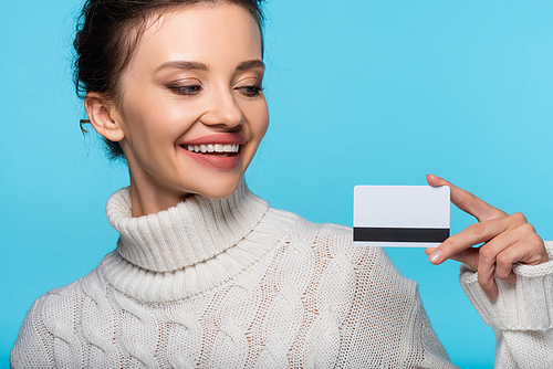 Smiling woman in knitted sweater looking at credit card isolated on blue