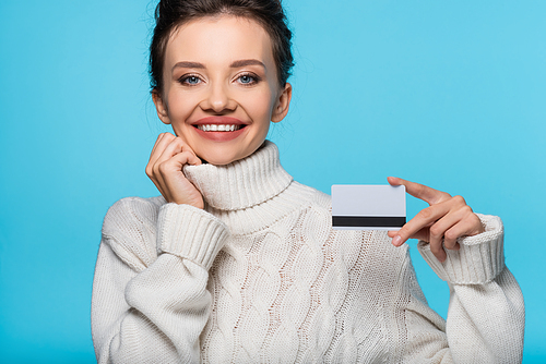 Positive woman in warm sweater holding credit card and  isolated on blue