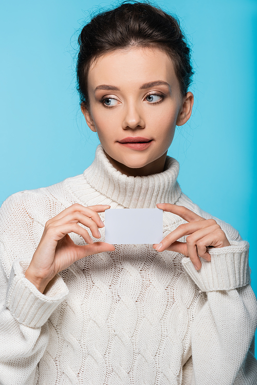 Pensive woman in sweater holding blank card isolated on blue
