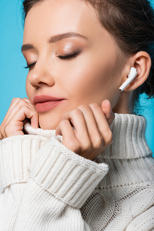 Portrait of woman with closed eyes wearing sweater and using earphone isolated on blue