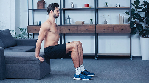 side view of muscular sportsman in shorts squatting near sofa at home