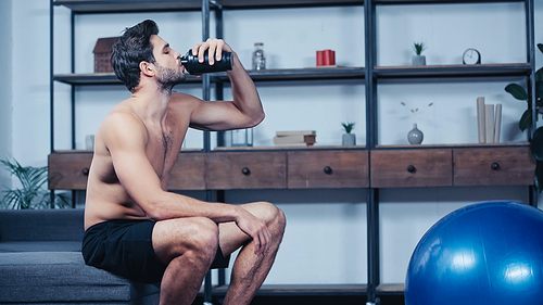 shirtless sportsman sitting on sofa and drinking water from sports bottle