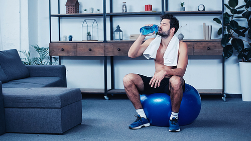 tired sportsman with towel sitting on fitness ball and drinking water from sports bottle