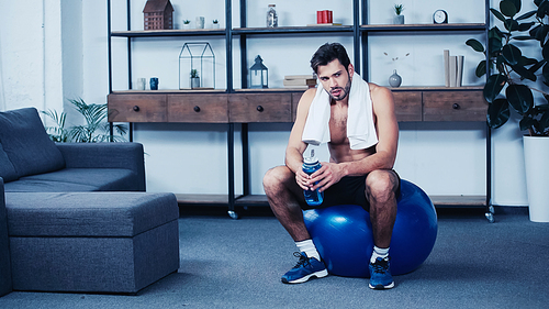 exhausted sportsman with towel sitting on fitness ball with sports bottle