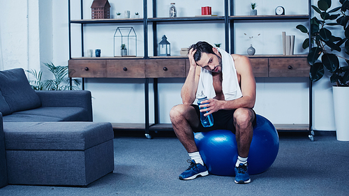 tired sportsman with towel sitting on fitness ball with sports bottle