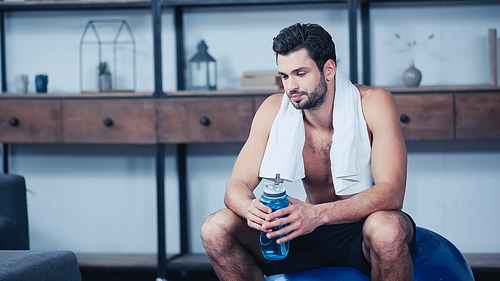 exhausted sportsman with towel sitting on fitness ball and holding sports bottle
