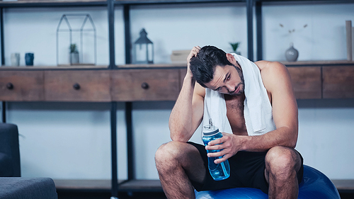 tired sportsman with towel sitting on fitness ball and holding sports bottle