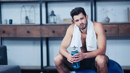 exhausted sportsman with towel sitting on fitness ball and holding sports bottle with water